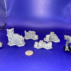 May include: A set of six gray 3D printed miniature buildings and a robot, all on a blue surface. The buildings are in various states of ruin, with some having collapsed walls and others having missing roofs. The robot is standing on a black base and has a detailed design.