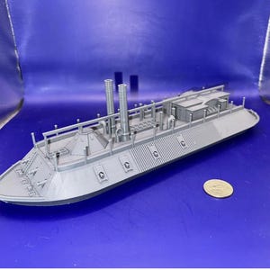 Może przedstawiać: Szary model statku z dwoma kominami, szczegółowym pokładem i monetą dla skali. Model ma długi, niski profil ze skomplikowanymi detalami wzdłuż boków i pokładu. Statek jest na niebieskim tle.