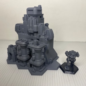 Puede incluir: Modelo gris impreso en 3D de una estructura de ciudad futurista con múltiples torres y detalles arquitectónicos. Una pequeña figura de robot gris se encuentra sobre una base hexagonal cercana. El modelo es probablemente para juegos de mesa o exhibición.
