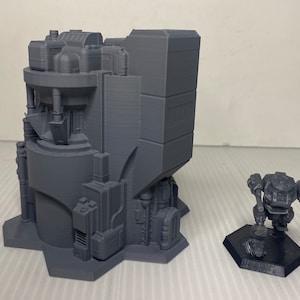 以下が含まれることがあります： 複雑なディテールを持つ未来的な建物の灰色の3Dプリントモデル。六角形の台座に小さなロボットフィギュアが隣接しています。建物は円筒形の上面と長方形の基部を持っています。