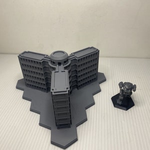 以下が含まれることがあります： 中央の塔と3つの翼を持つ、グレーの3Dプリントされた多層階建ての建物のモデル。小さなグレーのロボットフィギュアが、近くの黒い六角形のベースに立っています。建物モデルには多くの窓があります。