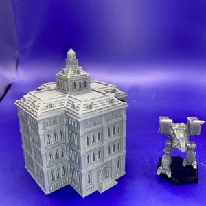 May include: A gray, 3D-printed model of a multi-story building with a detailed facade and a domed roof. Beside it is a gray mech robot model on a black base. The models are set against a blue background.