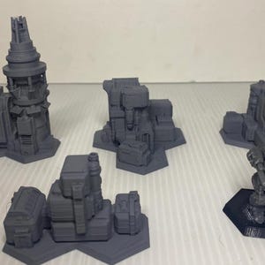 May include: A collection of gray, 3D-printed miniature terrain pieces, including buildings and a mech figure. The structures vary in design, with towers, rectangular blocks, and hexagonal bases. The pieces are designed for tabletop gaming.