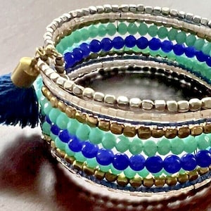 May include: A multi-strand bracelet with layers of blue, teal, and silver beads. The bracelet has a tassel with a gold-colored cap and a dark blue tassel. The bracelet is a wrap style.