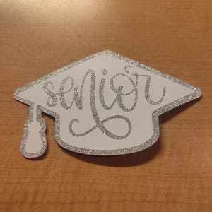 May include: A graduation cap-shaped cutout featuring a white background with a silver glitter border and the word senior written in silver glitter cursive script. A silver glitter tassel hangs from the left side of the cap.