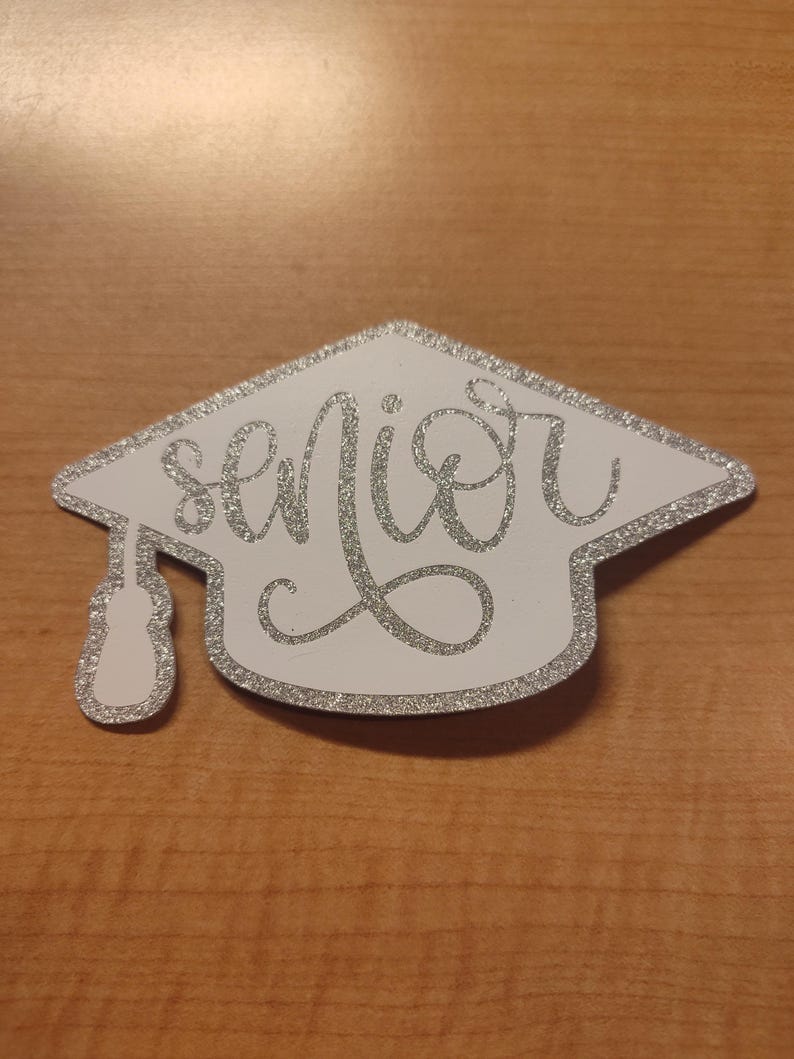 May include: A graduation cap-shaped cutout featuring a white background with a silver glitter border and the word senior written in silver glitter cursive script. A silver glitter tassel hangs from the left side of the cap.