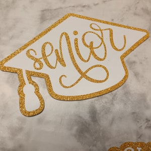 May include: A white graduation cap cutout with a gold glitter border and the word senior written in gold glitter cursive script across the center. The cap features a gold glitter tassel hanging from the left side, creating a decorative topper.