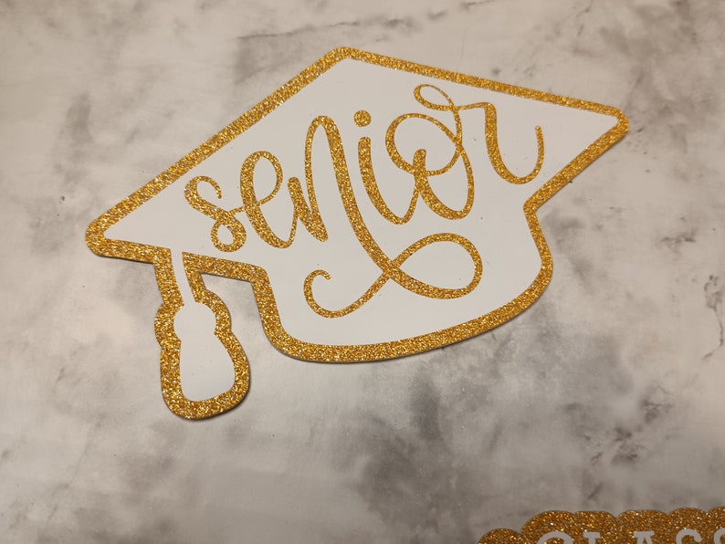 May include: A white graduation cap cutout with a gold glitter border and the word senior written in gold glitter cursive script across the center. The cap features a gold glitter tassel hanging from the left side, creating a decorative topper.