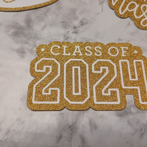 May include: A gold glitter cardstock cutout featuring the white text "CLASS OF 2024" in a bold, collegiate-style font. The cutout has a scalloped edge design and is positioned alongside other similar glittery paper decorations.