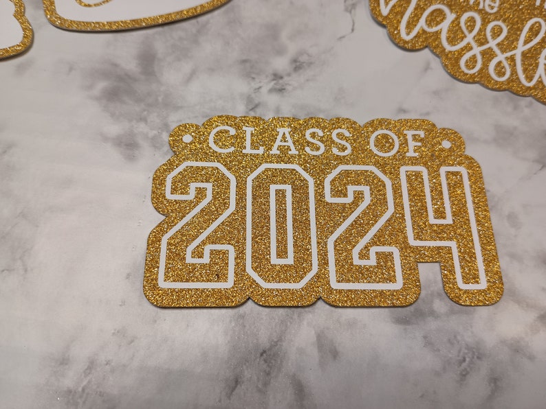 May include: A gold glitter cardstock cutout featuring the white text "CLASS OF 2024" in a bold, collegiate-style font. The cutout has a scalloped edge design and is positioned alongside other similar glittery paper decorations.