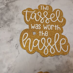 May include: A gold glitter cardstock cutout featuring the white text The tassel was worth the hassle in a decorative script and sans-serif font combination. The cutout has a scalloped edge shape and a smooth, reflective, sparkling surface texture.