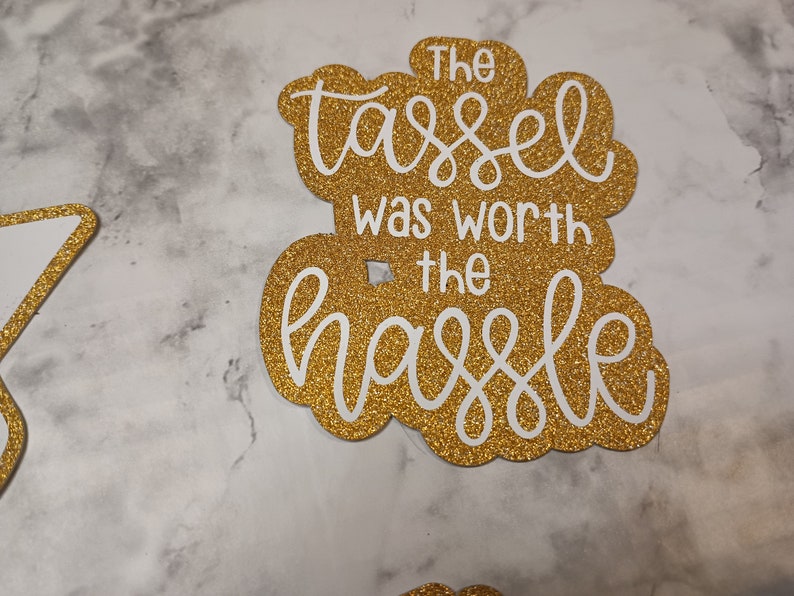 May include: A gold glitter cardstock cutout featuring the white text The tassel was worth the hassle in a decorative script and sans-serif font combination. The cutout has a scalloped edge shape and a smooth, reflective, sparkling surface texture.