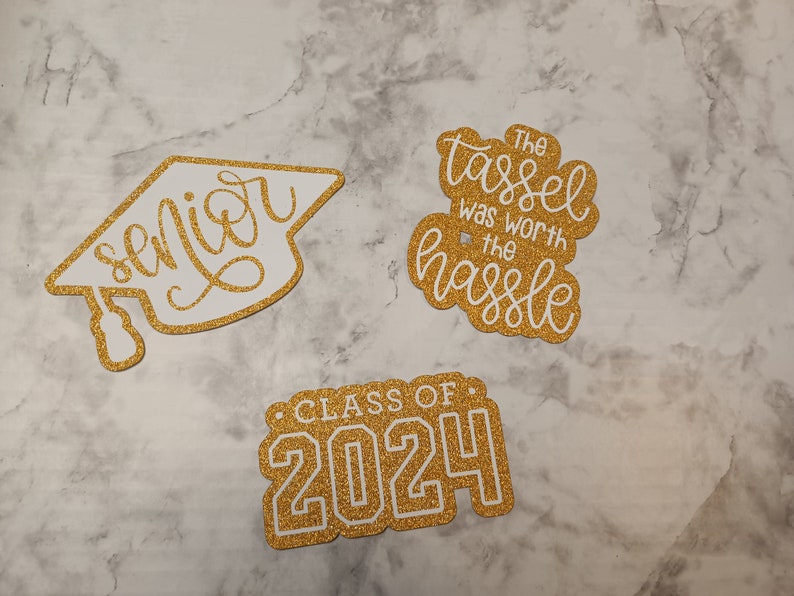 May include: Three gold glitter die-cut decorations for graduation. One is a mortarboard shape with the word senior, one is a cloud shape with the text The tassel was worth the hassle, and one is a rectangular shape with the text CLASS OF 2024.