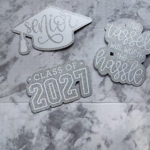 May include: Three silver glitter graduation-themed cutouts. One is a mortarboard shape with the word senior. Another is a rectangular shape with the text CLASS OF 2021. The third is a cloud-like shape with the text the tassel was worth the hassle.