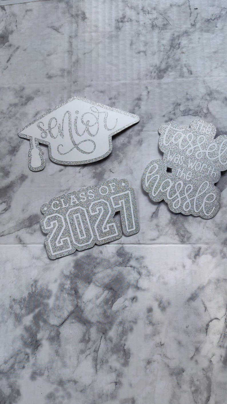 May include: Three silver glitter graduation-themed cutouts. One is a mortarboard shape with the word senior. Another is a rectangular shape with the text CLASS OF 2021. The third is a cloud-like shape with the text the tassel was worth the hassle.