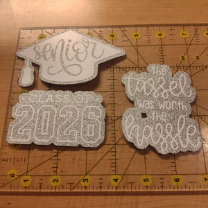 May include: Three silver glitter graduation-themed cutouts on a clear ruler. One is a graduation cap with the word "senior", one says "CLASS OF 2026", and one says "The tassel was worth the hassle". The ruler shows measurements in inches.