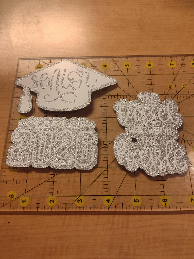 May include: Three silver glitter graduation-themed cutouts on a clear ruler. One is a graduation cap with the word "senior", one says "CLASS OF 2026", and one says "The tassel was worth the hassle". The ruler shows measurements in inches.
