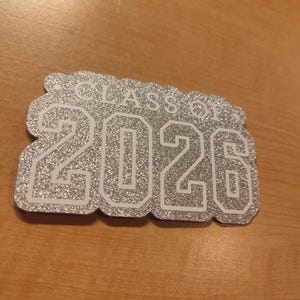 May include: A silver glittery cutout sign with a scalloped edge featuring the white text "CLASS OF" above the large white numbers "2026". The design has a textured, sparkling finish and a bold, block-style font for the year.