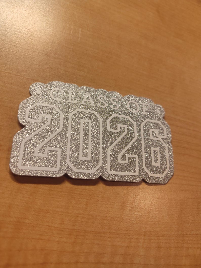 May include: A silver glittery cutout sign with a scalloped edge featuring the white text "CLASS OF" above the large white numbers "2026". The design has a textured, sparkling finish and a bold, block-style font for the year.
