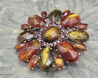 May include: A large, ornate brooch with a flower design. The brooch is made of metal and has a cluster of faceted, iridescent stones in shades of brown, gold, and green. The stones are set in a silver-toned metal and are surrounded by a ring of smaller, pink rhinestones.
