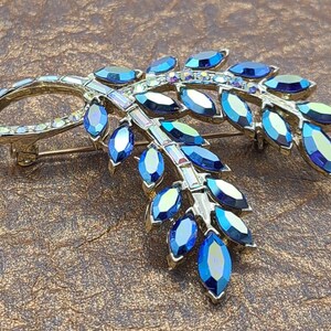 Könnte beinhalten: Eine goldfarbene Brosche mit einem Design aus blauen und schillernden Kristallen. Die Brosche ist in Form eines Zweigs mit Blättern gestaltet.