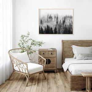May include: Black and white photograph of a foggy forest scene. The image is framed and hanging on a white wall above a wooden bed and nightstand.