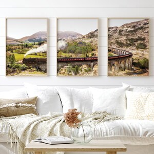 May include: A set of three framed prints featuring a scenic landscape with a train traveling over a stone bridge. The train is red and black, and the bridge is made of stone arches. The landscape is green and brown, with hills in the background.