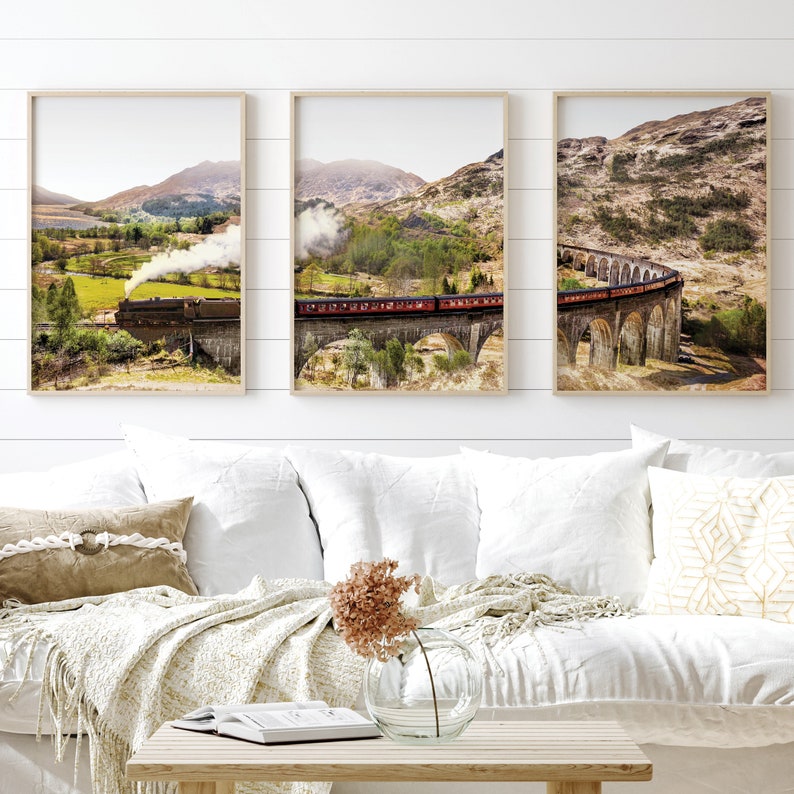 May include: A set of three framed prints featuring a scenic landscape with a train traveling over a stone bridge. The train is red and black, and the bridge is made of stone arches. The landscape is green and brown, with hills in the background.