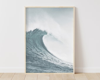 May include: A framed print featuring a large ocean wave. The wave is a mix of blue and white, with a foamy crest. The print is in a light wood frame, suitable for coastal or nautical decor.