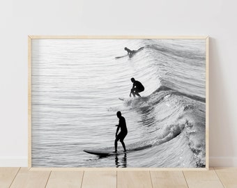 May include: Black and white photograph of three surfers riding waves. The surfers are silhouetted against the bright sky and water. The waves are crashing and breaking, creating a dynamic scene.