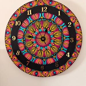 May include: A circular black wall clock with a detailed mandala pattern. The clock has gold numerals and hands. The mandala design is composed of pink, yellow, teal, and orange, creating a vibrant and decorative piece for the wall.