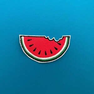 May include: A watermelon-shaped patch with a bite taken out. The patch features a red interior with black seeds, a white border, and a green rind. The patch is set against a solid blue background.