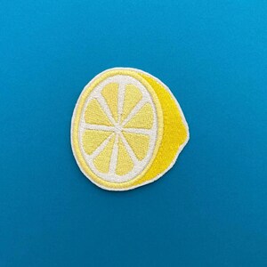 May include: Embroidered patch featuring a yellow lemon slice with a white outline and details. The patch is set against a solid blue background. The lemon slice design is a bright, cheerful image.