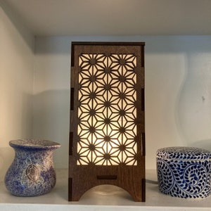 May include: A wooden lamp with a geometric pattern cut-out design. The lamp is lit and casts a warm glow through the pattern. The lamp is on a shelf with two decorative ceramic pieces.