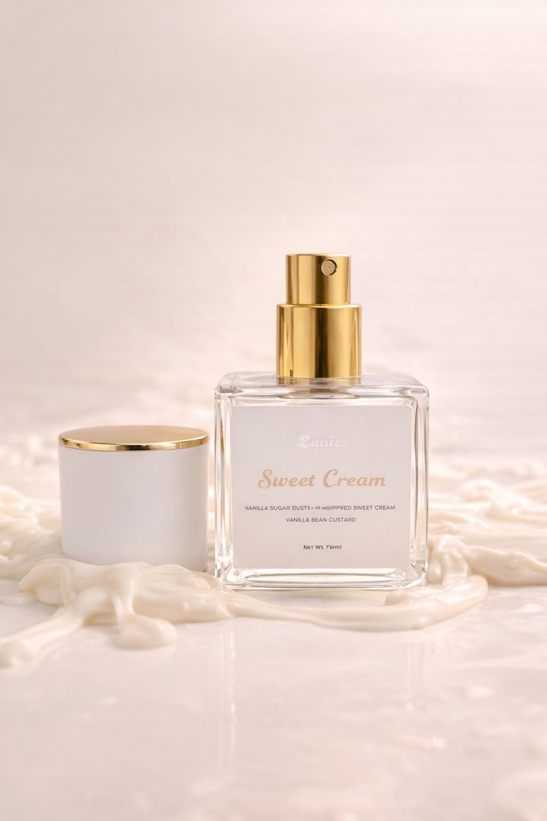 May include: A clear glass perfume bottle with a gold cap and a white label that reads "Sweet Cream." The bottle is next to a white cap with a gold rim. The text on the label also says "Vanilla Sugar Dust - Whipped Sweet Cream" and "Vanilla Bean Custard."