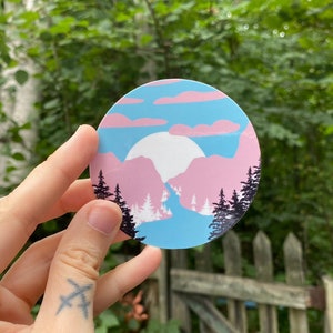 Puede incluir: Una pegatina redonda con un diseño de paisaje de montaña rosa y azul. La pegatina presenta una luna blanca y una silueta de árboles en primer plano.