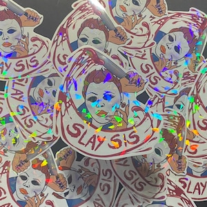 May include: A set of holographic stickers featuring a masked figure holding a knife and the text "SLAY SIS". The stickers are iridescent and have a glittery finish.