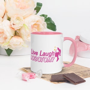 May include: White ceramic mug with a pink rim and handle. The mug has a pink graphic of a woman's head with a brain and stars, and the text "Live Laugh Lobotomy".