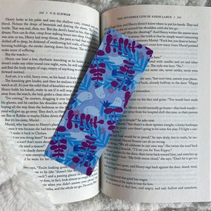 May include: A close-up of a purple and blue bookmark with a floral pattern and a ghost design. The bookmark is placed on a page of a book with text about a character named Henry.