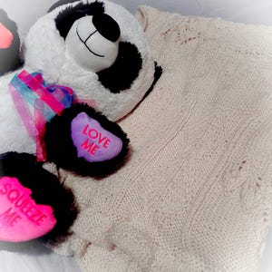 May include: A white and black panda plush toy with pink and blue ribbon. The panda is holding two heart-shaped plush toys that say "Love Me" and "Squeeze Me". The panda is lying on a cream-colored knitted blanket.
