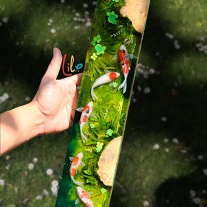 Puede incluir: Una pieza de arte rectangular de resina con peces koi nadando en un entorno verde y acuoso con plantas verdes y detalles de madera. Los peces koi tienen marcas rojas y naranjas. La pieza se sostiene en una mano.