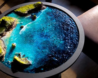 May include: Round coffee table with a detailed ocean scene. The tabletop features a miniature landscape with green mossy islands and a vibrant blue ocean. The table has a dark gray frame and a dark base.