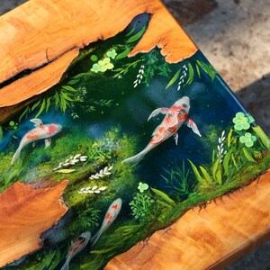 May include: Wooden table with a resin inlay showing a koi pond scene. The table has a natural wood grain border and a clear resin top with painted koi carp, aquatic plants, and a dark blue water effect.