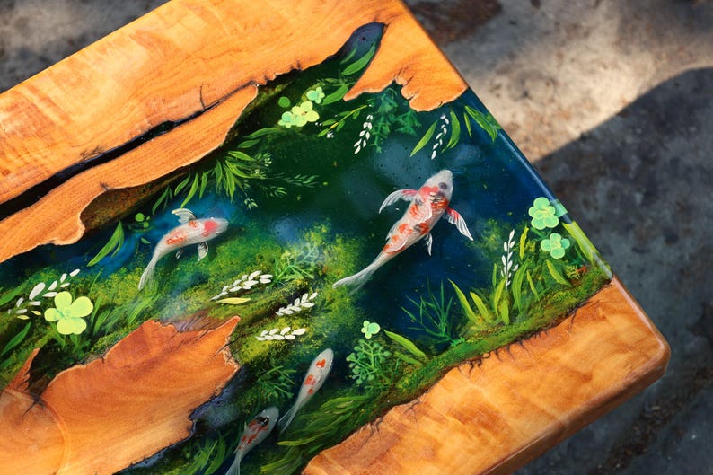 May include: Wooden table with a resin inlay showing a koi pond scene. The table has a natural wood grain border and a clear resin top with painted koi carp, aquatic plants, and a dark blue water effect.