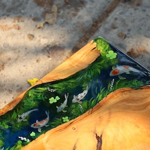May include: Wooden serving tray with a river design. The tray features a clear resin river with koi fish, green plants, and clover accents. The tray's wooden surface has a natural wood grain.