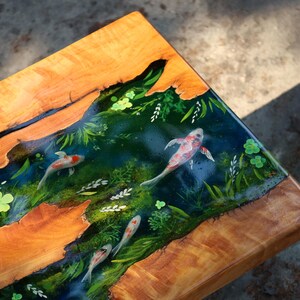 May include: A wooden table with a resin inlay showing a koi pond scene. The table has a natural wood grain border and a central design of koi fish swimming amongst painted aquatic plants. The koi are red, white, and black.