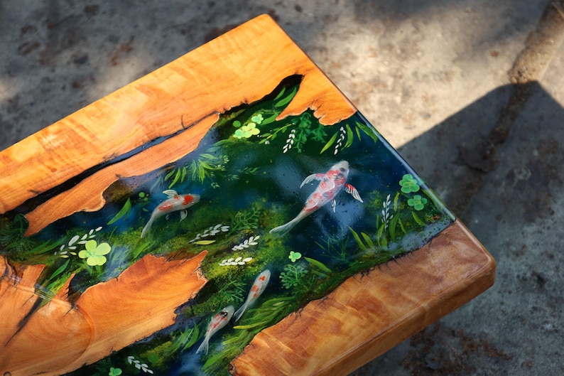 May include: A wooden table with a resin inlay showing a koi pond scene. The table has a natural wood grain border and a central design of koi fish swimming amongst painted aquatic plants. The koi are red, white, and black.