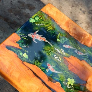 May include: Wooden table with a resin top depicting a koi pond. The resin is a deep blue, with painted koi carp and aquatic plants. The table's frame is made of natural wood with a warm, orange hue.
