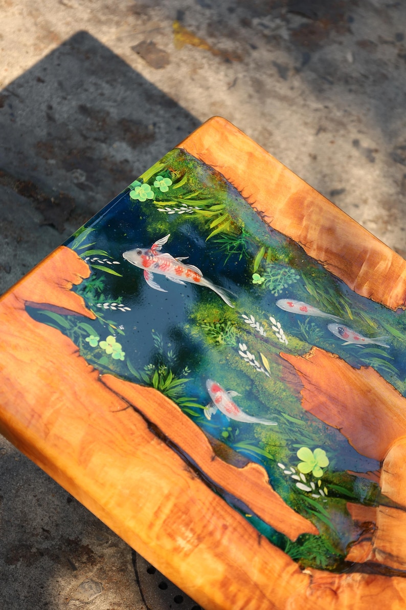 May include: Wooden table with a resin top depicting a koi pond. The resin is a deep blue, with painted koi carp and aquatic plants. The table's frame is made of natural wood with a warm, orange hue.