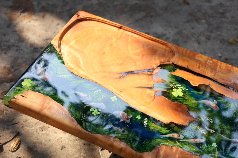 May include: Wooden serving tray with a river design. The tray has a light brown wooden border and a central river scene with koi carp, green plants, and blue water. The river is made of resin.
